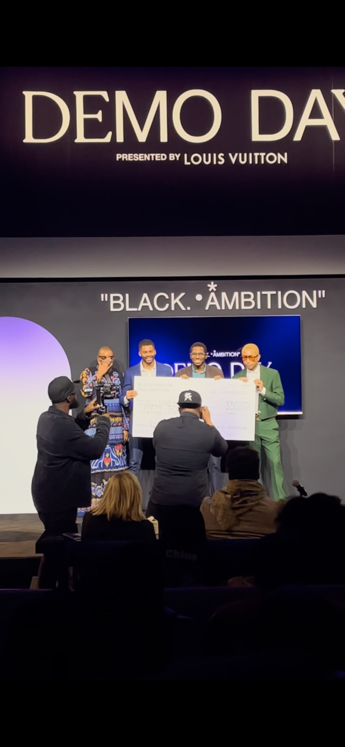 Demo Day presented by Louis Vuitton — Black Ambition founders holding an oversized prize check on stage with Pharrell Williams